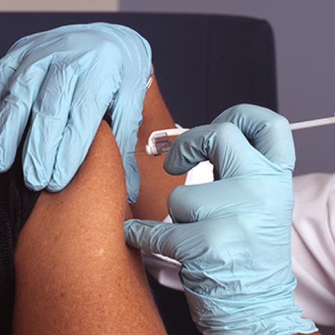 A nurse wearing blue gloves administers a vaccine (vaccination) into a male patient's arm.