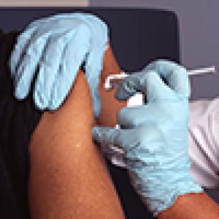 A nurse wearing blue gloves administers a vaccine (vaccination) into a male patient's arm.