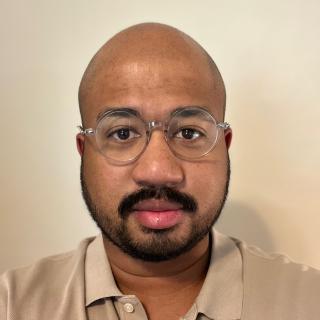 Headshot of Timothy Watts. Mr. Watts is a black man. He is wearing a tan shirt and glasses with clear frames. He has a beard and mustache.